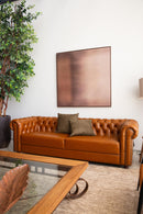 Sofá Chesterfield em couro Amelie caramelo com encosto em capitonê, dimensões de 250 cm de largura, 75 cm de altura e 80 cm de profundidade. Estrutura de madeira tratada, espuma certificada e percintas elásticas italianas. Ideal para ambientes sofisticados. Outros itens na fotografia não estão incluídos.