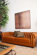 Sofá Chesterfield em couro Amelie caramelo com encosto em capitonê, dimensões de 250 cm de largura, 75 cm de altura e 80 cm de profundidade. Estrutura de madeira tratada, espuma certificada e percintas elásticas italianas. Ideal para ambientes sofisticados. Outros itens na fotografia não estão incluídos.